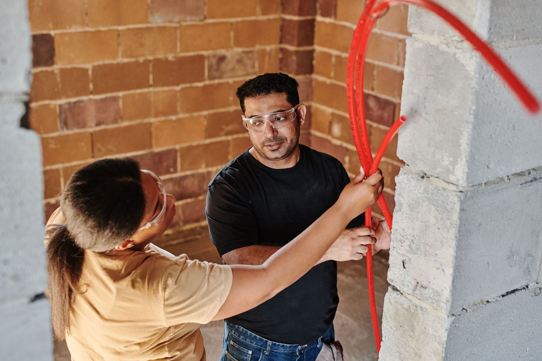 Two workers guiding red tubing through a wall opening at a job site